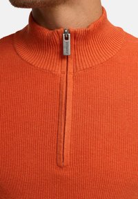 Orange, struktureret halv-zip trøje med ribbet krave. Har en sølvfarvet lynlås med branding. Tætsiddende design.