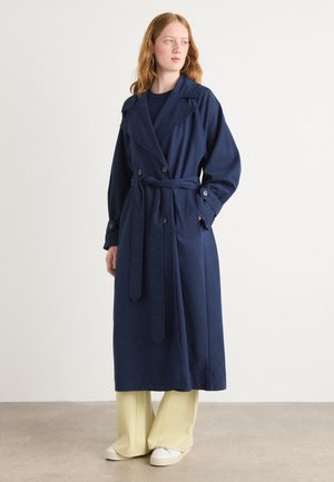 Marineblauer Trenchcoat mit einem Bindegürtel, großem Reverskragen und Knopfakzenten, kombiniert mit hellgelben weit geschnittenen Hosen und weißen Sneakers.