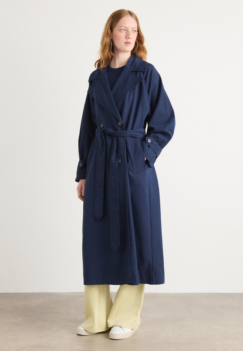 Manteau trench bleu marine avec une ceinture à nouer, grand col à revers et accents de boutons, associé à un pantalon large jaune clair et des baskets blanches.