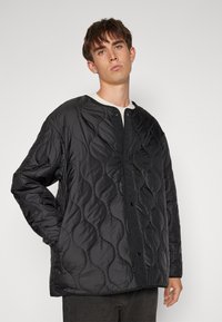 Black quilted jacket with a rounded neckline, snap button closure, and textured wave pattern. Made of lightweight fabric.