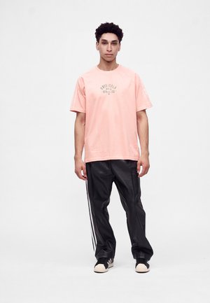 Joven de pie con una camiseta rosa con el texto "Originals Athletics", pantalones deportivos negros con rayas blancas y zapatillas negras y blancas.