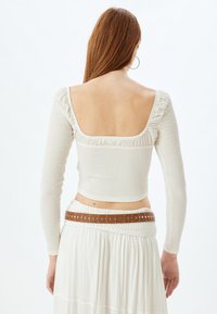 Witte, geribde lange mouw top met een vierkante halslijn en plooidetails langs de rand, gecombineerd met een witte rok en een bruine riem.