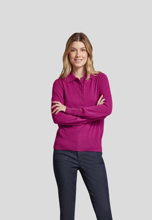 Fuchsianer Pullover mit Kragen, langen Ärmeln und Knopfdetaillierung an den Manschetten, kombiniert mit dunklen Jeans, vor einem grauen Hintergrund.