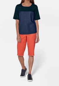 Maglietta a maniche corte color bloc in blu navy e blu scuro con taschino sul petto, abbinata a pantaloncini arancioni fino al ginocchio e scarpe da ginnastica scure.