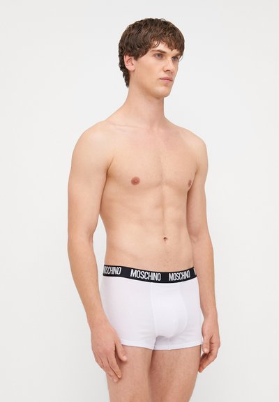 Biele bavlnené boxerky s čiernym pásom s logom Moschino, priliehavý strih a hladká textúra. Model zobrazený zboku.
