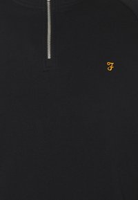 Black zip-up sweater with a metal zipper and small yellow embroidered logo on the left chest. Made from a smooth, textured fabric.