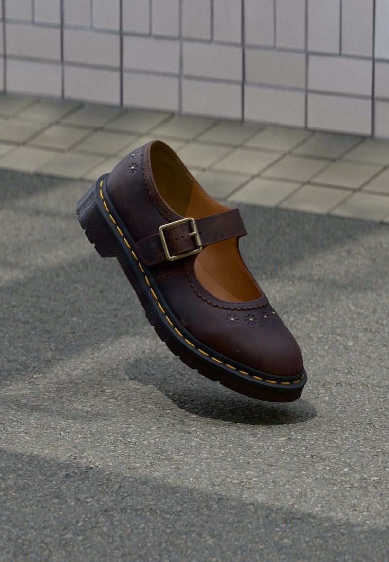 Chaussure Mary Jane en cuir marron foncé avec surpiqûres jaunes et boucle, flottant au-dessus d’un bitume texturé près d’un trottoir carrelé.