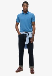 Chemise polo bleue avec un col et un logo brodé, assortie à un jean foncé avec les manches retroussées, complétée par des chaussures marron et une chemise bleu clair nouée autour de la taille.