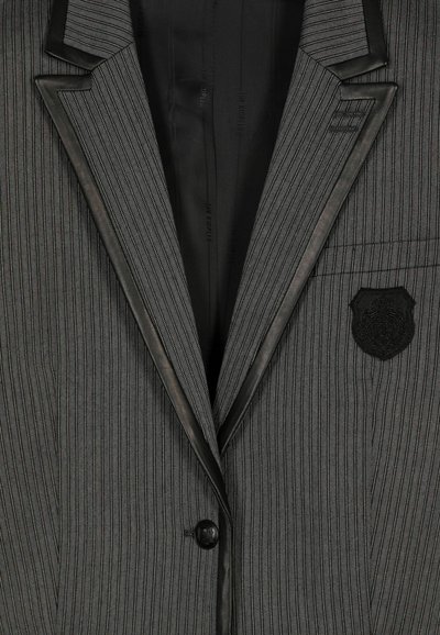 Grijs gestreepte blazer met zwarte satijnen revers, knoopsluiting en een borstzak met een zwart geborduurd embleem. Soepele textuur.
