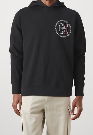 Mężczyzna w czarnej bluzie z kapturem z okrągłym logo "Hilfiger H Columbus Avenue Since 1985" na piersi, w zestawie z beżowymi spodniami typu cargo.