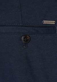 Temno modra tkanina z zapiranjem na gumb in majhno kovinsko značko z napisom "JACK & JONES." Gladek tekstura z nežnimi detajli šivanja.