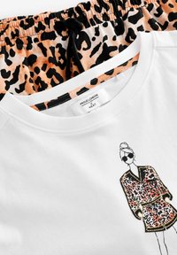 Biała bluza z kapturem z postacią z kreskówki w leopardzim wzorze. Poniżej, wzorzyste spodenki dresowe w kolorze pomarańczowym i czarnym, z elastycznym pasem.