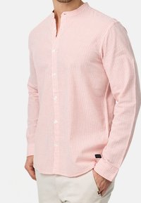 Camicia a maniche lunghe con colletto, di colore rosa chiaro con strisce verticali bianche, dotata di bottoni sul davanti e di una piccola etichetta sul lato inferiore.