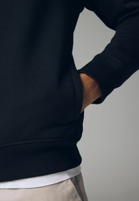 Sweat-shirt noir avec des poignets et un ourlet côtelés, comportant une poche latérale. Le tissu est doux, avec une texture lisse et une coupe décontractée.