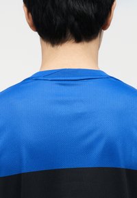 Πίσω όψη ενός ατόμου με κοντά μαύρα μαλλιά που φοράει μπλε και μαύρο αθλητικό πουκάμισο με υφή, πάνω σε ένα απλό φόντο.