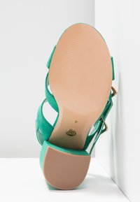 Sandale à talon haut en daim vert avec plusieurs brides, un talon fermé et une semelle en caoutchouc beige. Présente des détails en matériel doré.