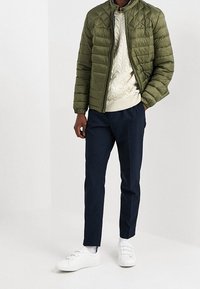 Giubbotto trapuntato verde oliva, maglione a trecce color crema, pantaloni blu navy e sneakers bianche, con un'enfasi sulla texture e sul contrasto dei colori.