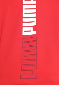 Červené tričko s názvem značky "PUMA" ve velkých bílých a tmavě modrých block písmenech, s hladkou bavlněnou texturou.