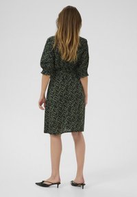 Robe verte avec un motif noir de feuillages, manches bouffantes et taille cintrée. L'ourlet tombe juste au-dessus du genou. Portée avec des mules à talons noirs.