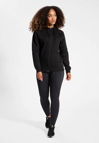 Sudadera con capucha negra con cremallera frontal, cordones ajustables y puños acanalados, combinada con leggings negros y zapatillas deportivas.