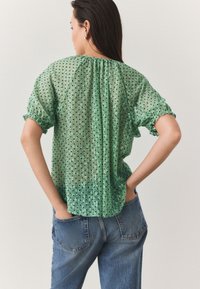 Green sheer blouse with short puff sleeves, dotted texture, and gathered neckline. Worn with high-waisted blue denim jeans.