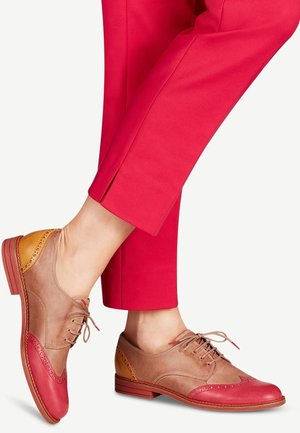 Tofarvede oxfordsko i brun, pink og sennepsgul båret med korte, lyse pink bukser mod en ensfarvet lys baggrund.
