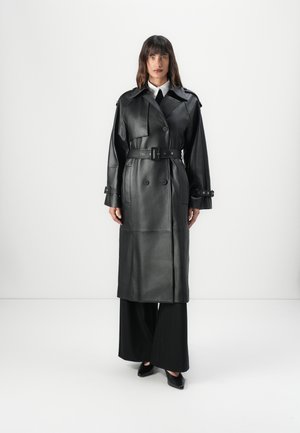 Cappotto trench in pelle nera con bottoni doppi, vita con cintura e ampi rever, abbinato a pantaloni neri a gamba larga e scarpe a punta.