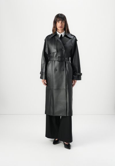 Manteau en cuir noir avec des boutons double boutonnage, une taille ceinturée et des revers larges, associé à un pantalon noir à jambes larges et des chaussures à bouts pointus.