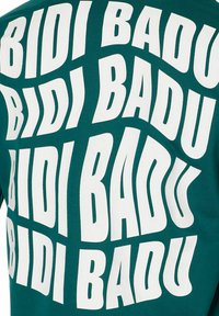 T-shirt in tessuto verde con la frase "BIDI BADU" in grande, grassetto, tipografia bianca disposta in varie orientamenti sulla superficie.