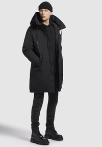 Melna parka ar augstu apkakli, ar noņemamu kapuci, ar rāvējslēdzēja aizdari un sānu kabatām, apvienota ar tumšiem džinsiem un melniem zābakiem.