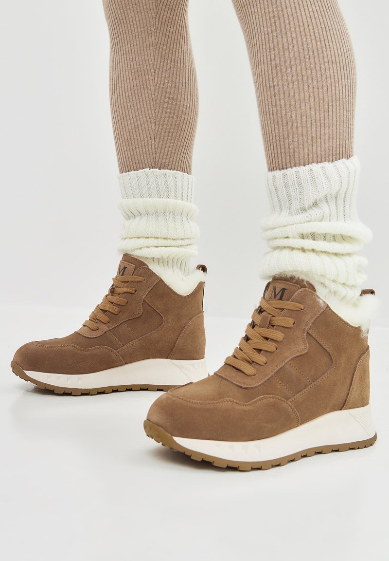 Bruine suède hoge sneakers met witte nepbont voering, getextureerde rubberen zool en vetersluiting. Gecombineerd met crèmekleurige geribbelde beenwarmers.