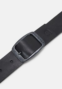 Svart läderbälte med en borstad metallspänne, som har en rektangulär form och en texturerad yta. Inkluderar flera justeringshål.