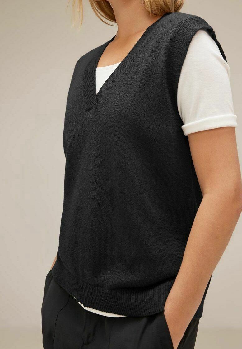 Black knitted vest with a V-neck, short sleeves, and ribbed hem, layered over a white T-shirt. Smooth texture and relaxed fit.
