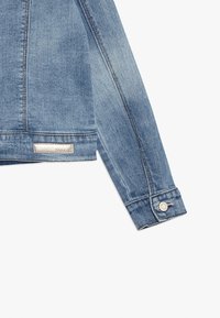 Světle modrá džínová bunda s mírně vybledlou texturou, s knoflíkovými manžetami a stříbrnou štítkou na lemu. Obsahuje klasický límec a detaily prošívání.