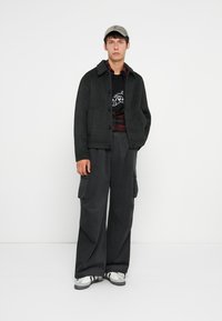 Donkergrijze, gestructureerde jas met knoopsluiting, gelaagd over een zwart T-shirt met graphic en een geblokt shirt, gecombineerd met losse zwarte cargo-broeken en sneakers.