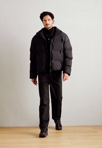 Doudoune noire avec fermeture éclair à l'avant et coupe oversize, associée à un pantalon noir et des chaussures noires. Texture lisse avec une finition mate.