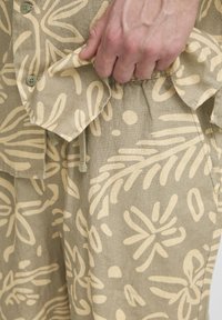 Helebeeži särg lillemustriga, valmistatud tekstuurse kangaga, millel on suured nööbid ja tasku detail, osaliselt hoitud käe poolt.