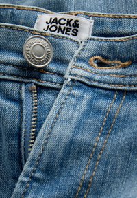 Gros plan sur un jean en denim bleu avec bouton en métal, fermeture éclair et étiquette en tissu blanc portant la mention "JACK&JONES" au-dessus de la braguette.