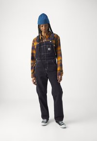 Mørke denim overalls med justerbare stropper, en brystlomme, kombinert med en rutete oransje-og-blå flanellskjorte og en blå strikket lue. Sorte sneaker.