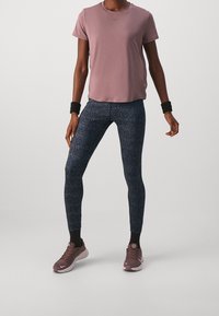 Kobieta w fioletowej koszulce z krótkim rękawem, czarnych legginsach wzorzystych, fioletowych tenisówkach, czarnych skarpetkach i czarnych opaskach na nadgarstkach stoi na jednolitym tle.