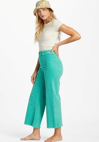 Teal corduroy brede benbukser med frynset kant, høj talje og forlommer, parret med en lys beige tætsiddende t-shirt og en mønstret bøttehat.