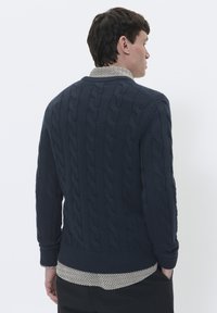 Maglione blu navy a trecce con orlo e polsini a costine, indossato sopra una camicia a fantasia con colletto rotondo.
