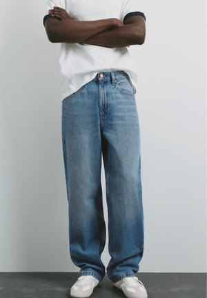 Person med mørk hud iført en hvid t-shirt, løse blå jeans og hvide sneakers, står med armene krydset mod en ensfarvet baggrund.