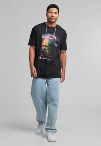 Sort bomulds T-shirt med et farverigt kraniet grafik og tekst på forsiden, parret med lyseblå denimjeans og hvide sneakers.