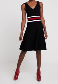 Robe en maille noire avec un col en V, ornée d'une rayure horizontale rouge et blanche à la taille. Jupe évasée portée avec des bottines rouges.