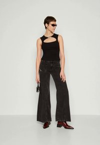Top preto sem mangas com um design recortado, combinado com jeans pretos de perna larga e botas de tornozelo borgonha. Segura uma pequena mala preta.