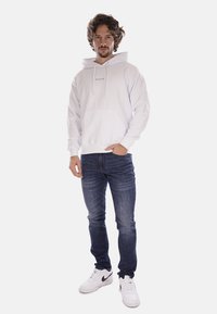 Felpa bianca in cotone con tasca frontale e lacci, abbinata a jeans blu scuro e sneakers bianche con dettaglio swoosh nero.