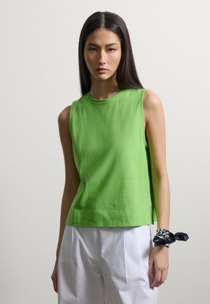 Femme aux longs cheveux foncés portant un haut vert vif sans manches, un pantalon blanc et une écharpe noire à motifs nouée autour de son poignet.