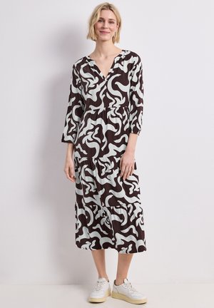 Femme debout portant une robe midi à motif abstrait noir et blanc avec des manches trois-quarts et des baskets blanches.