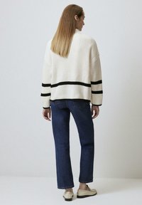 Un pull en tricot blanc avec des rayures noires sur les manches, au design ample, associé à un jean droit bleu foncé et à des chaussures de couleur claire.